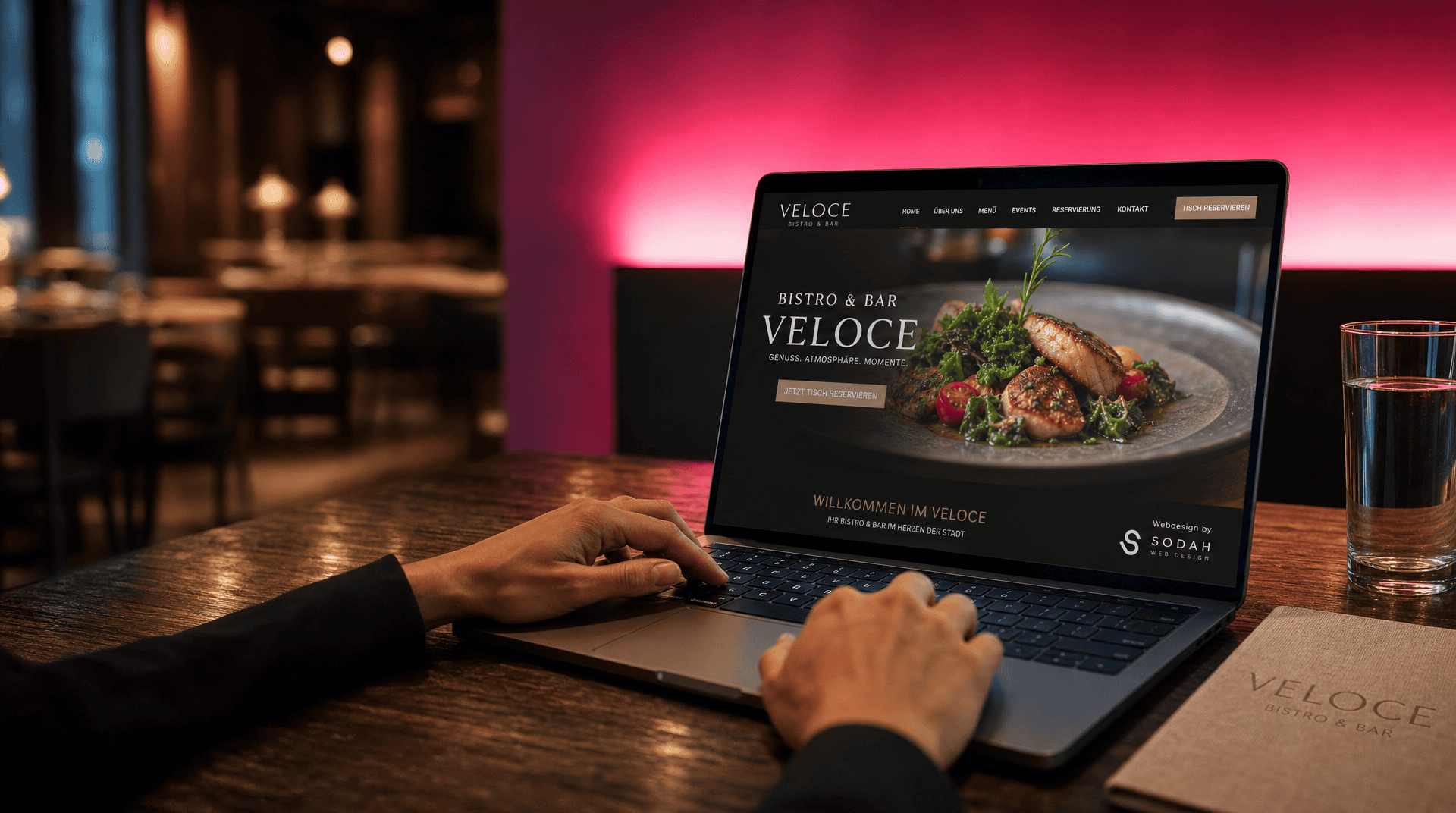 Person bedient Laptop mit Restaurant-Website (VELOCE Bistro & Bar), die ein appetitliches Gericht zeigt. Im Hintergrund ein Restaurantambiente mit Menü und Wasserglas.